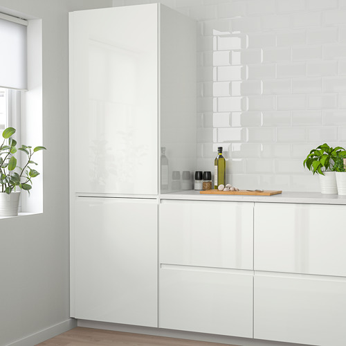 Una moderna cocina blanca con gabinetes VOXTORP brillantes y protector contra salpicaduras. Las plantas en macetas y la botella de aceite de oliva se colocan en la encimera.