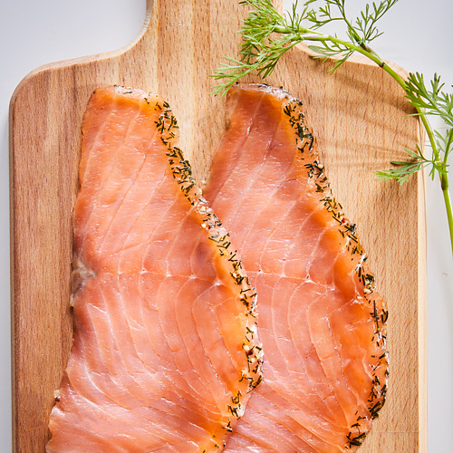 Tabla de cortar de madera con dos filetes de salmón ahumado de color naranja rosado, adornados con eneldo.