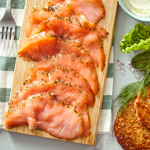 Salmón ahumado en frío SJÖRAPPORT exhibido en una tabla de madera con eneldo fresco, lechuga, cebollas y galletas saladas.
