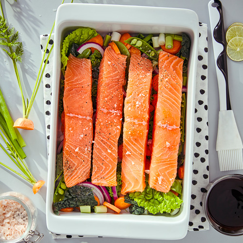 Bandeja blanca con filetes de salmón y verduras coloridas sazonadas con sal y pimienta, lista para cocinar o servir.