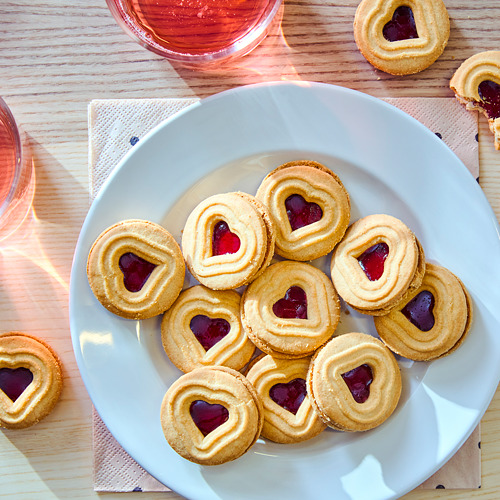 Plato de galletas rellenas de mermelada en forma de corazón, de color marrón dorado, con centros de gelatina de color rojo rosado y vasos de líquido rosado al lado.