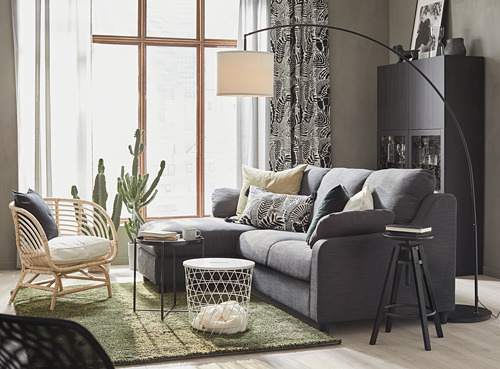 Moderno salón con lámpara de pie SKAFTET gris, sofá, silla de mimbre, cactus y armario negro.