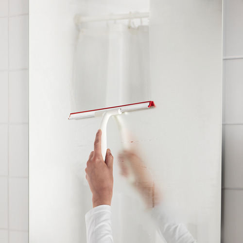 Una persona está limpiando la pared de una ducha con una escobilla de goma blanca con mango rojo, eliminando el agua y manteniendo la limpieza.