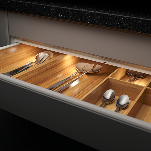 Cajón de cocina abierto con compartimentos de madera, que contiene varios utensilios metálicos. La iluminación LED cálida incorporada ilumina el contenido.