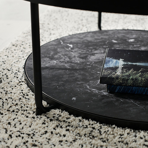 Mesa auxiliar redonda negra con tapa de cristal oscuro, patas metálicas y un libro sobre ella, sobre una alfombra de manchas.