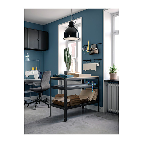 Oficina en casa con paredes azules, un banco de trabajo de metal negro, mesa de madera, silla de oficina gris, máquina de coser, plantas en macetas y una lámpara colgante negra.