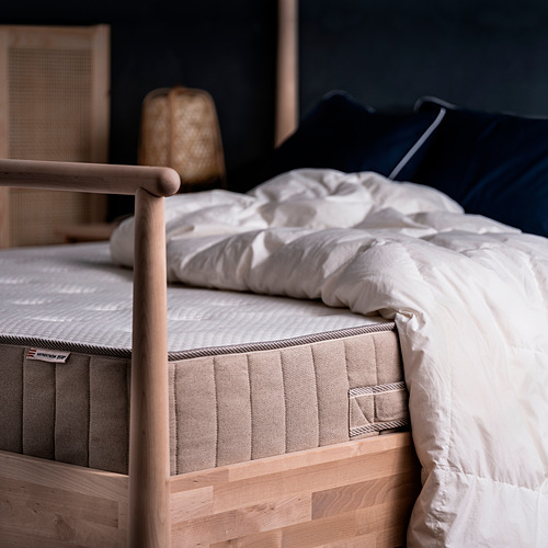 Un acogedor dormitorio con una cama de madera marrón claro con un colchón VATNESTRÖM blanco, almohadas azul marino y un cabecero de lino beige.