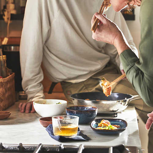 Dos personas cocinando con la sartén IKEA 365+, usando palillos. Comida en sartén, tazón y un vaso en la mesa.