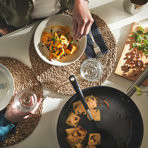 Una persona que sirve comida cocinada de una sartén en un plato. La sartén se coloca sobre una mesa con manteles individuales y vasos.