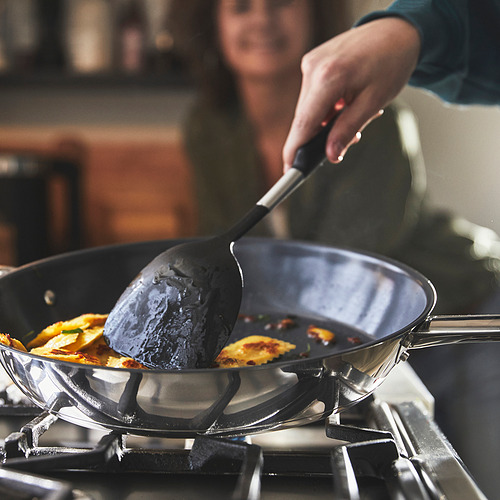 Una persona cocina en una sartén grande antiadherente IKEA 365+ en una estufa, revolviendo trozos de tortilla amarilla con una espátula. La sartén tiene dos asas para facilitar la elevación.