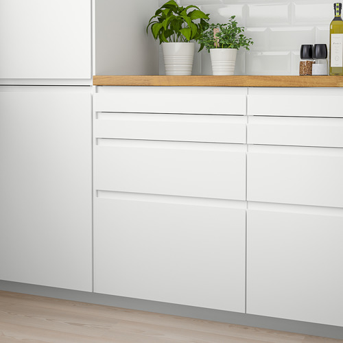 Cocina blanca moderna con tiradores elegantes. Las pequeñas plantas en macetas se sientan en el mostrador. Los armarios son de acabado mate, altos y estrechos, con tiradores integrados.