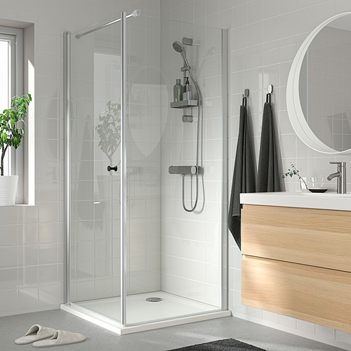 Baño moderno con ducha de vidrio, toallas oscuras, tocador de madera, azulejos blancos y grises, espejo redondo, planta en maceta.