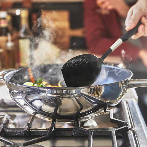 Cocina en curso: una mano revuelve las verduras en un wok en una estufa de gas. El vapor se eleva de la sartén, lo que indica una cocción activa.