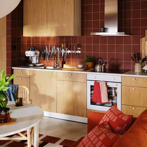 Cocina con gabinetes de madera clara, placa para salpicaduras de azulejos rojos y un acogedor sofá con almohadas a cuadros. El paño de cocina RINNIG de algodón rojo cuelga perfectamente en el mango del horno.