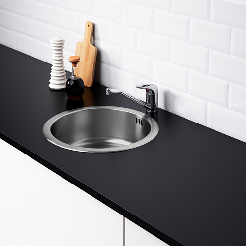 Fregadero de acero inoxidable, grifo cromado, encimera negra, azulejos blancos, tabla de cortar, condimentos.