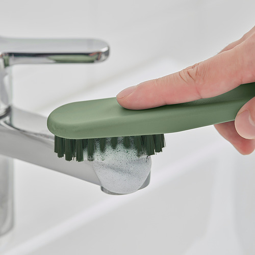 Persona que usa un cepillo verde con cerdas densas para limpiar alrededor del grifo de la cocina o el baño.