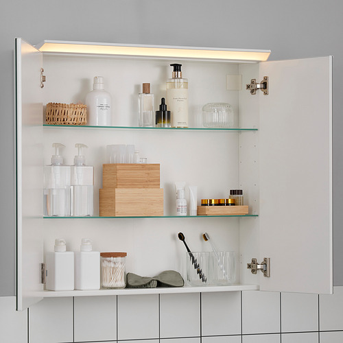 Gabinete de baño abierto con puertas ligeras y espejadas que revelan estantes de vidrio llenos de artículos de tocador, cajas de madera y artículos de cuidado personal.