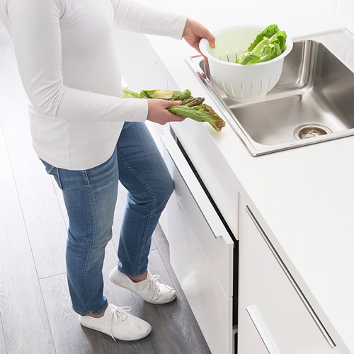 Una persona con camisa blanca y jeans usa un cajón de la cocina con un abridor de empuje, sosteniendo lechuga verde sobre un fregadero.