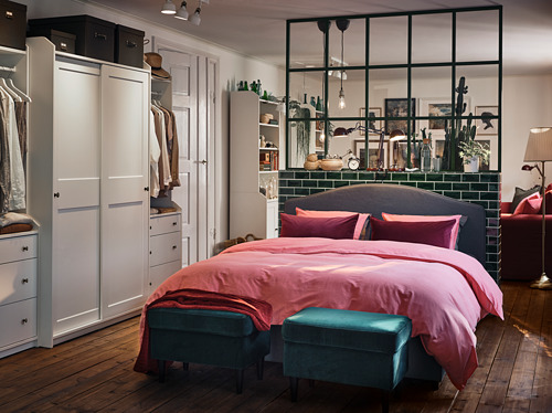 Armarios HAUGA de dormitorio moderno en blanco, ropa de cama rosa, otomanas azules y zona de cama dividida en cristal.