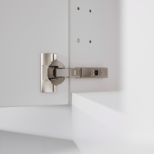 Una bisagra de cocina moderna, ajustable en tres direcciones, montada sobre un mueble blanco. Ideal para una alineación precisa de la puerta y una fácil extracción para la limpieza.