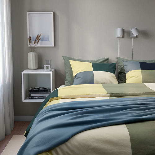 Cama con edredón de cuadros terrosos BRUNKRISSLA, almohadas, mesita de noche con jarrón, agua, reloj, libros.