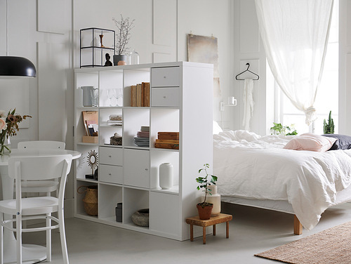 Dormitorio minimalista con estantes blancos KALLAX, que exhiben libros, jarrones y decoración. Una cama con sábanas blancas y una planta en maceta cerca.
