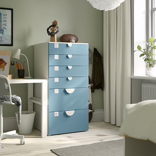 Cómoda SMÅSTAD azul de 5 cajones en el dormitorio, entre el escritorio y la ventana, que contiene pequeños artículos de decoración.