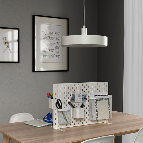 SKÅDIS Separador de escritorio blanco con bolsillos sobre escritorio de madera. Bolígrafos, tijeras, cuaderno. Dos piezas de arte mural. Luz central.