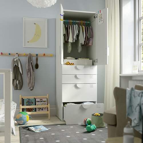 Habitación infantil con armario SMÅSTAD blanco, expositor abierto de ropa y cajones. Juguetes coloridos en el suelo, la silla y la estantería con mantas colgantes.