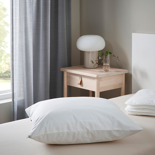Dormitorio minimalista: cama blanca con almohadas grandes, mesita de noche, lámpara y jarrón. Ventana con cortinas.
