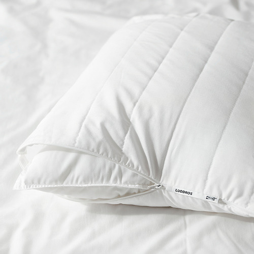 Primer plano de un protector de almohada LUDDROS blanco con cremallera visible, colocado sobre una cama.