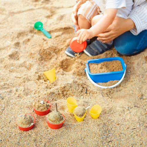Niños jugando con juguetes de arena SANDIG, incluyendo tazas y una cuchara, desarrollando habilidades motoras en la arena.