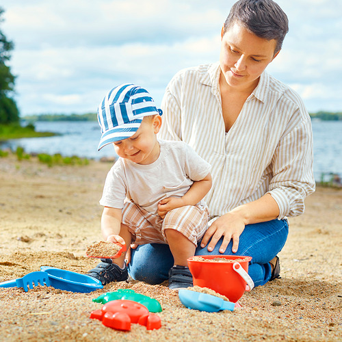 Una mujer y un niño juegan con juguetes de arena en la playa. Utilizan un cubo, un rastrillo y una pala, lo que mejora las habilidades motoras y la creatividad.