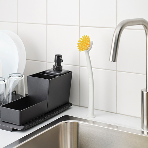 Un área de fregadero de cocina con un dispensador de jabón gris, un cepillo de cerdas amarillas con un mango blanco unido a la pared y un grifo de acero inoxidable.