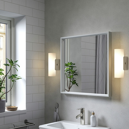 Baño moderno con gran espejo NISSEDAL flanqueado por luces. Fregadero blanco, planta verde, azulejos grises. El espejo refleja el espacio, añadiendo profundidad.