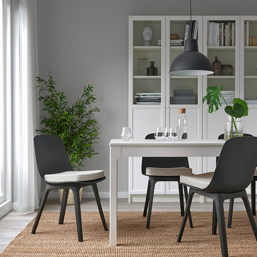 Comedor moderno con paredes blancas, sillas grises, mesa blanca, lámpara negra, plantas y libros.