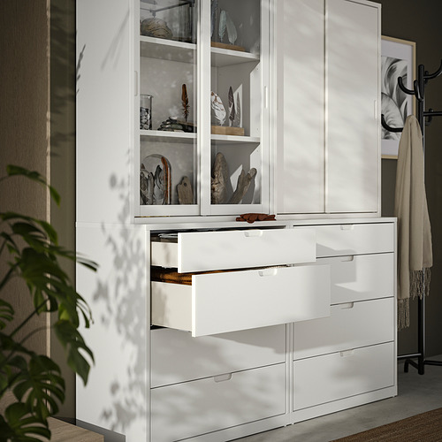 Mueble y cómoda TONSTAD blanco con puertas de cristal y tiradores de madera, donde se exponen elementos decorativos, junto a un toallero y una planta.
