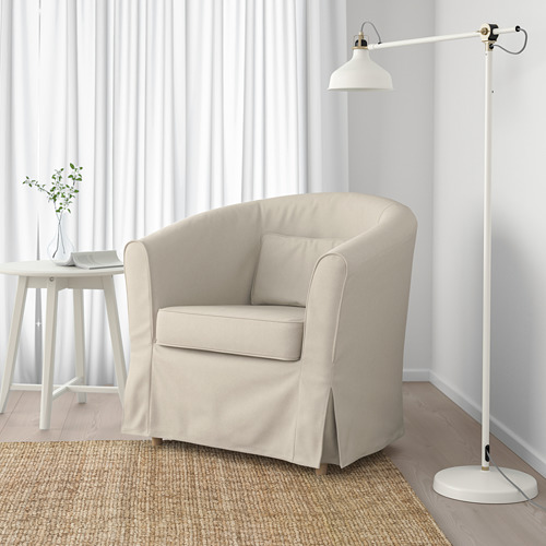 Un acogedor sillón beige con un cojín se encuentra sobre una alfombra tejida cerca de una lámpara de pie blanca alta. Detrás, una mesa auxiliar blanca sostiene una planta y un libro.