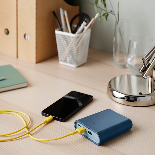 Banco de energía azul en el escritorio, teléfono de carga con cable amarillo. Más pequeño que la taza, junto a un bolígrafo, un cuaderno verde y una lámpara.