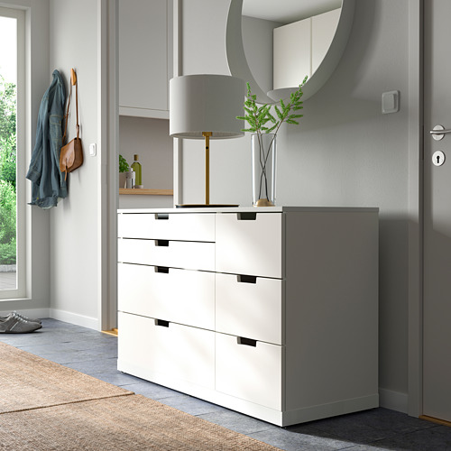 Pasillo moderno con cofre NORDLI blanco, lámpara, espejo y percheros. La cómoda tiene cuatro cajones, colocados contra una pared cerca de una puerta.