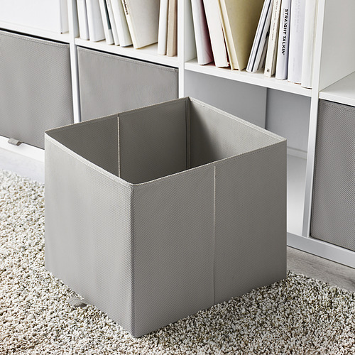 Caja de almacenamiento plegable gris vacía junto a la estantería sobre el suelo alfombrado.