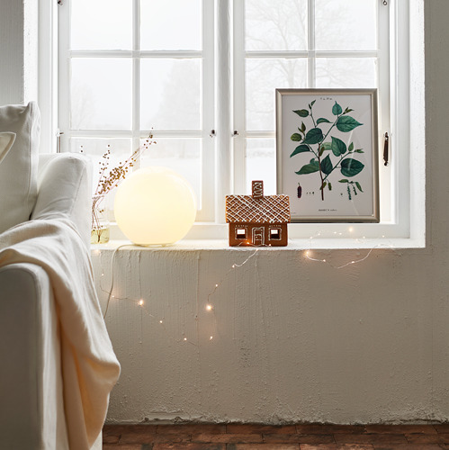 Acogedora sala de estar con una gran lámpara de globo blanco en el alféizar de la ventana, decorada con luces de hadas y una pequeña casa de pan de jengibre.