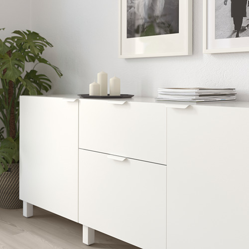 Mueble blanco con asas minimalistas, exhibiendo velas y revistas apiladas en la parte superior, junto a una planta.