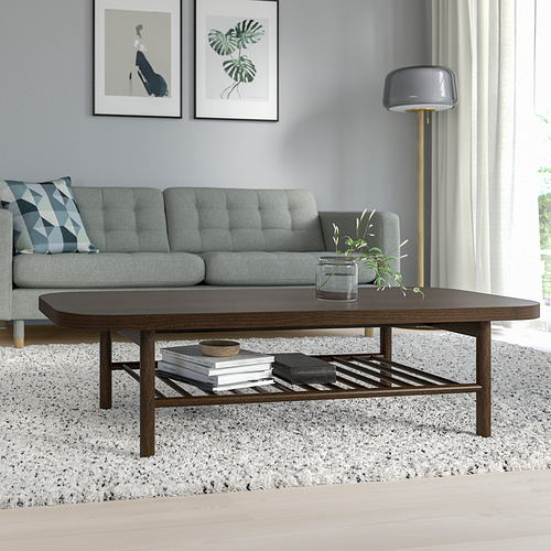 Sala de estar moderna con mesa de centro de madera marrón oscuro LISTERBY, sofá gris y lámpara de pie alta. La mesa tiene un estante inferior con libros y una planta.