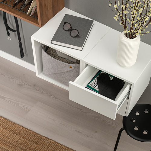 Mueble de almacenaje EKET blanco con dos compartimentos abiertos, uno que contiene un cubo de fieltro gris y el otro un cajón negro con libros y guantes a la vista.