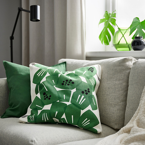 Cojín Underbloomma en verde/blanco sobre sofá gris con almohadas verdes, lámpara y planta.