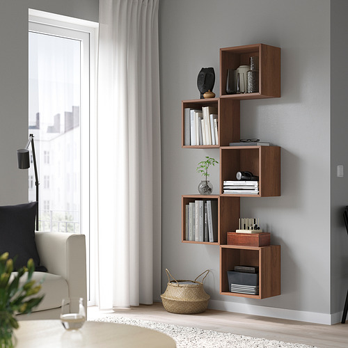 Estanterías de pared EKET en color nogal, que contienen libros y decoración, colocadas junto a una ventana y un sofá. Su diseño modular maximiza el espacio en la pared.
