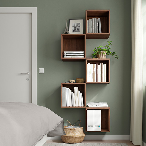 Dormitorio moderno con estantes asimétricos EKET de color marrón oscuro montados en la pared, que contienen libros, decoración y una planta, junto a una cama blanca y una puerta blanca cerrada.