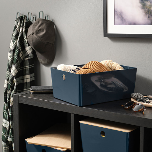 Caja de almacenamiento azul oscuro con tapa de madera llena de ropa en una estantería oscura junto a gafas de sol y bufanda.