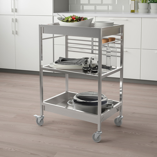 Carro de cocina KUNGSFORS plateado con 3 estantes, sosteniendo platos, vasos y ollas, que encaja perfectamente en una cocina moderna con gabinetes blancos.